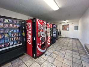 Fila de màquines expenedores amb el logotip de Coca-Cola, un sòl de rajoles i una paret sense decoració amb una porta.