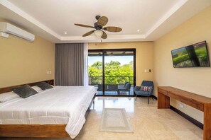 Ein Schlafzimmer mit Bett, Deckenventilator, Fernseher, einem Balkon mit Stuhl und Blick ins Grüne.