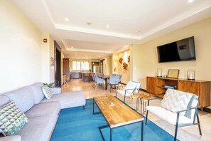 A modern living room with a sofa, coffee table, and chairs. There is a dining area with a table and chairs in the background. A TV is mounted on the wall. - Condo in Playa Ocotal | Sunset Terrace & Pool (Playa Ocotal)