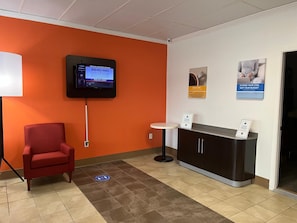 Ein Empfangsbereich mit einem roten Sessel, einem digitalen Bildschirm, einem kleinen runden Tisch und Informationsplakaten an der Wand.