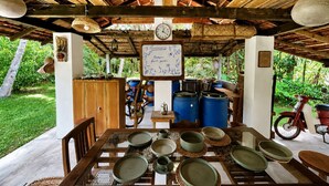 Uma zona de refeições rústica, com uma mesa de madeira posta para uma refeição, rodeada por diversos utensílios e equipamentos de cozinha.