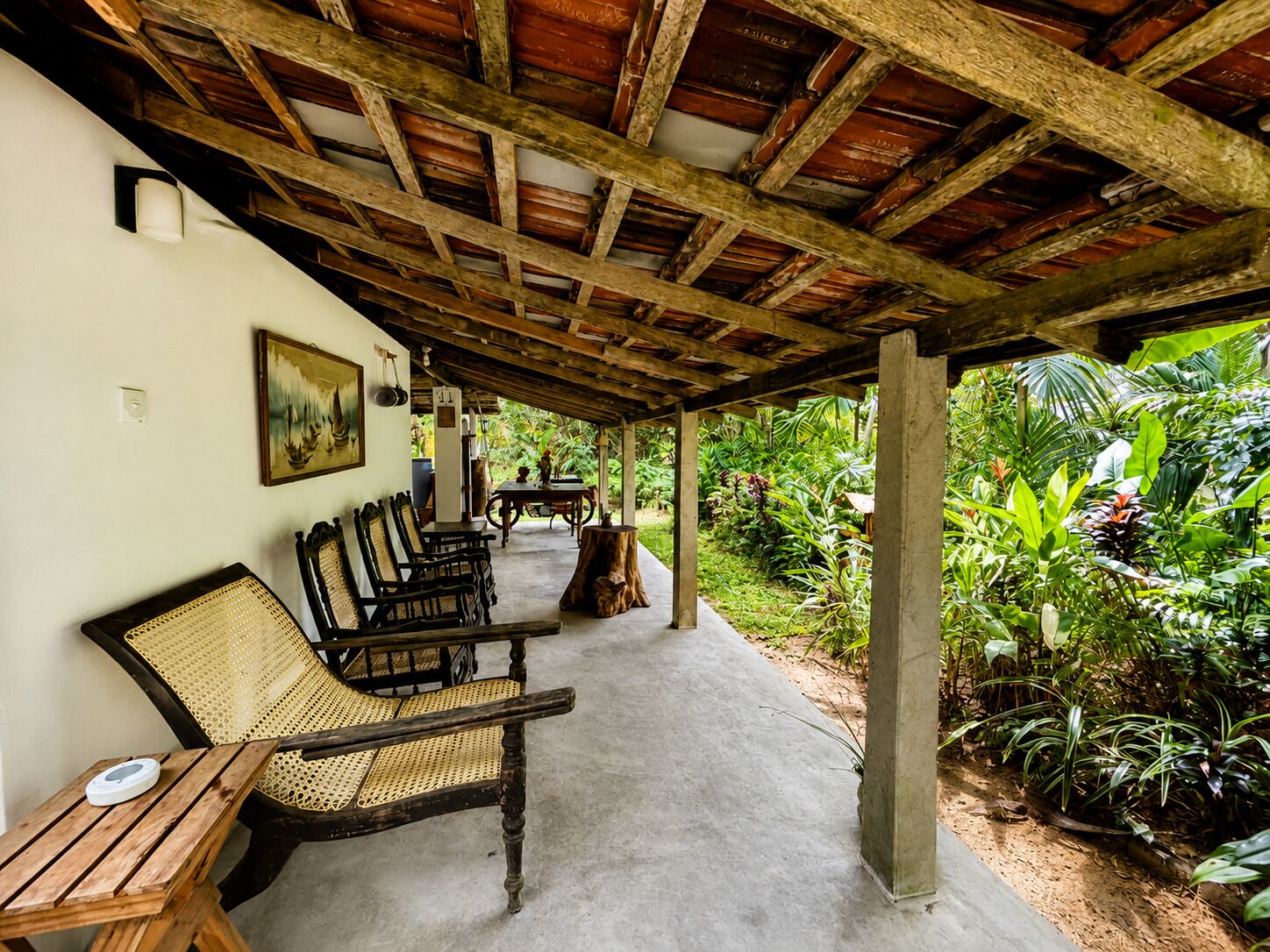 An outdoor covered seating area with rattan chairs and a table, surrounded by lush greenery.