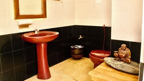 Um banheiro com uma pia vermelha, uma lixeira preta pequena, um vaso sanitário e uma estátua em um banquinho de madeira.