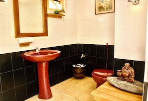 A bathroom with a red sink, a small black trash bin, a toilet, and a statue on a wooden stool.
