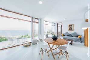A modern living room with a large window, a round wooden table, and a grey sofa with cushions.