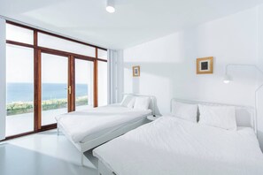 A modern bedroom with two beds, large windows, and a view of the sea.