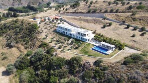 A modern two-story building with a swimming pool, surrounded by a landscaped garden and a hilly landscape.