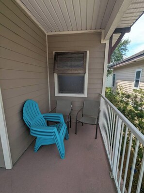 A covered porch with two chairs and a table, a window with blinds, and a white railing. - Luna Del Mar - Beach Oasis Home - Pool/Relax/Grill (Destin)