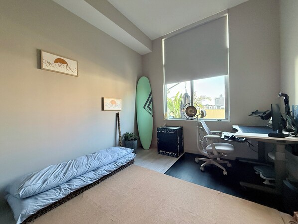 Un dormitorio con una cama, un escritorio con una computadora, una tabla de surf y una ventana con vista a edificios.
