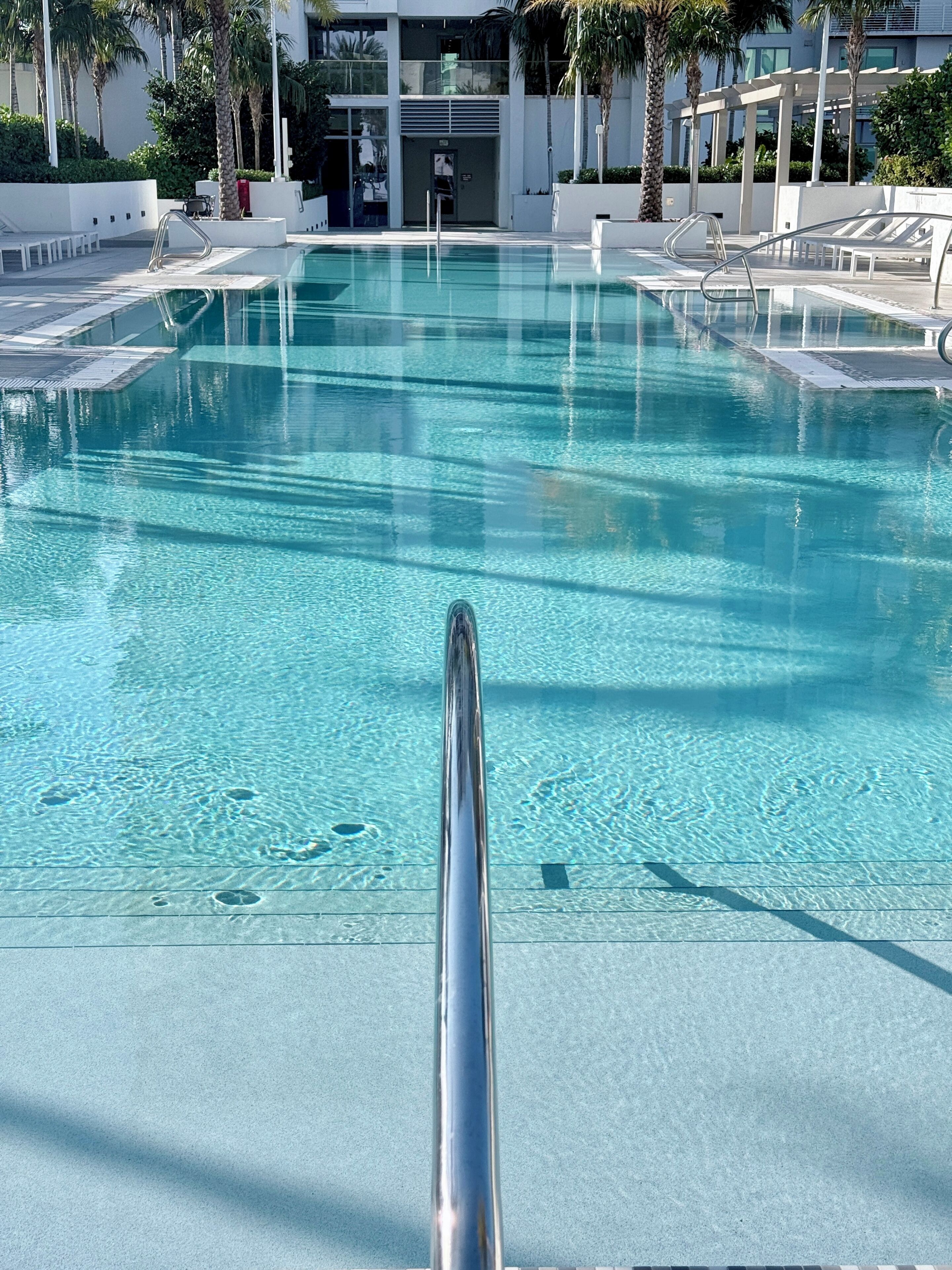 Une piscine aux eaux cristallines, avec une rambarde métallique en premier plan, entourée de chaises longues et de palmiers.