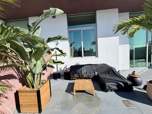Un espace extérieur aménagé, abrité et doté d’un canapé et d’une petite table, entouré de plantes en pot et d’une grande fenêtre.