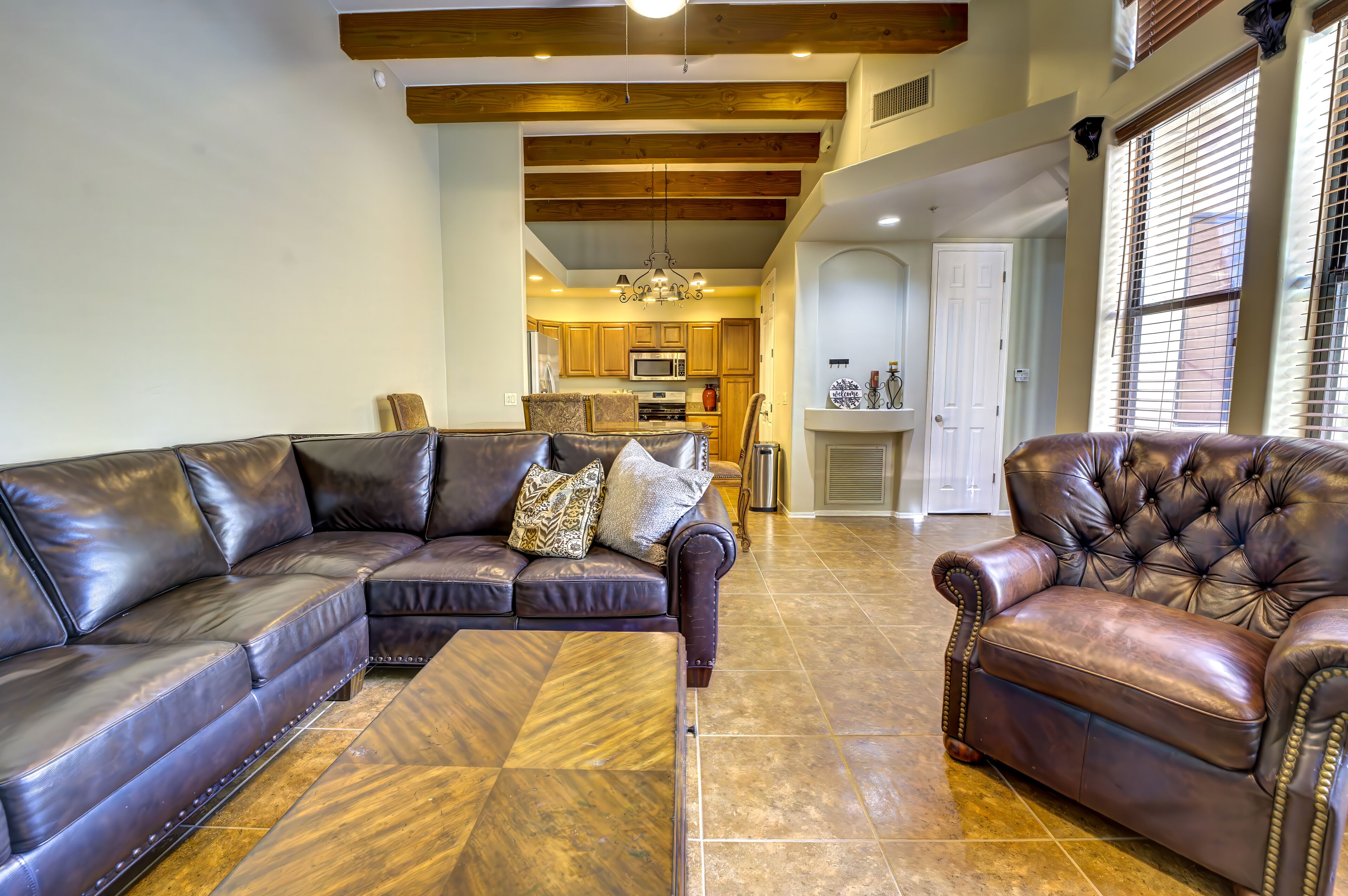 A spacious living room with a sectional sofa and a tufted armchair, a kitchen in the background, and a hallway leading to a door.