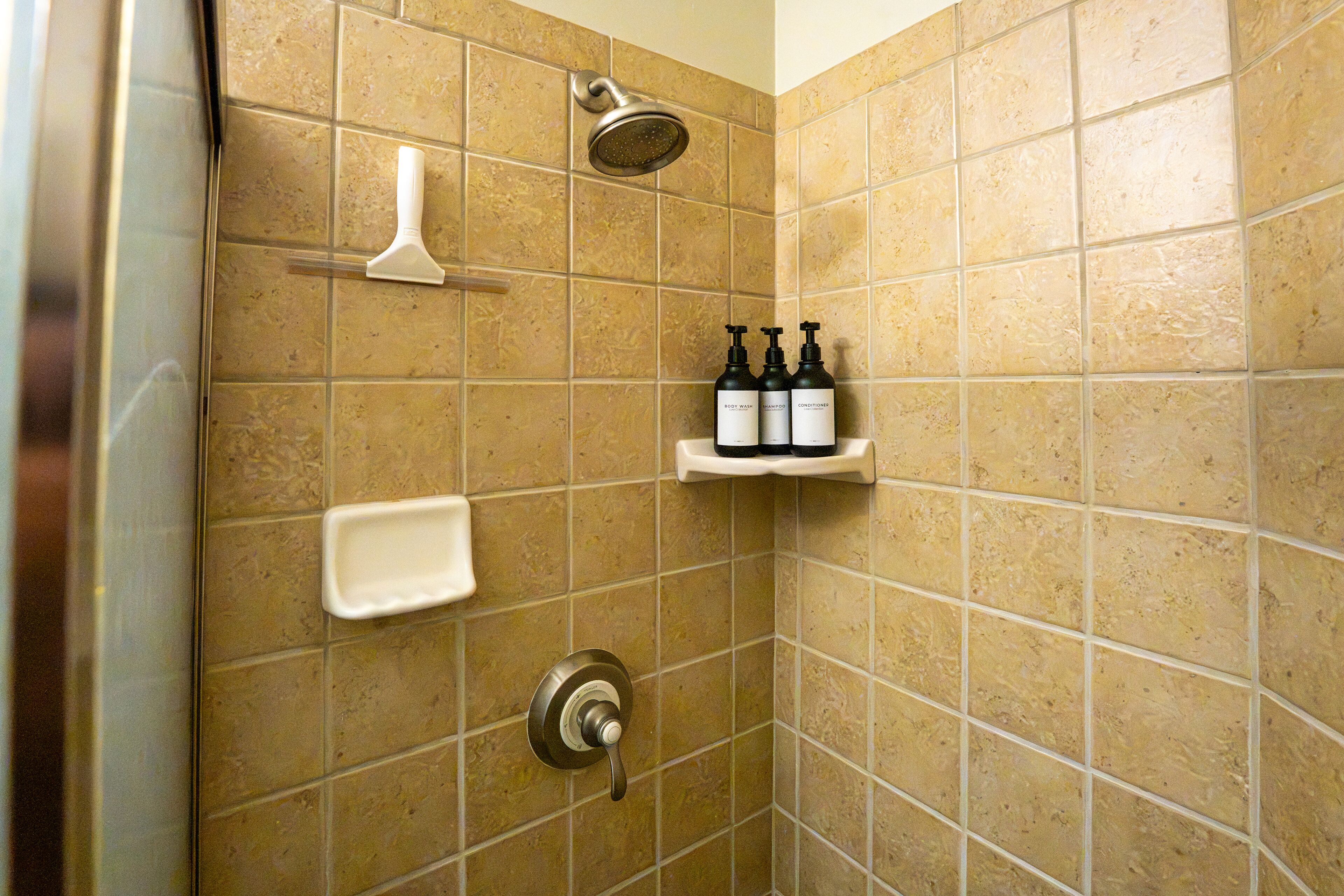 A shower area with a handheld showerhead, a handheld soap dispenser, and a shelf with three bottles of toiletries.