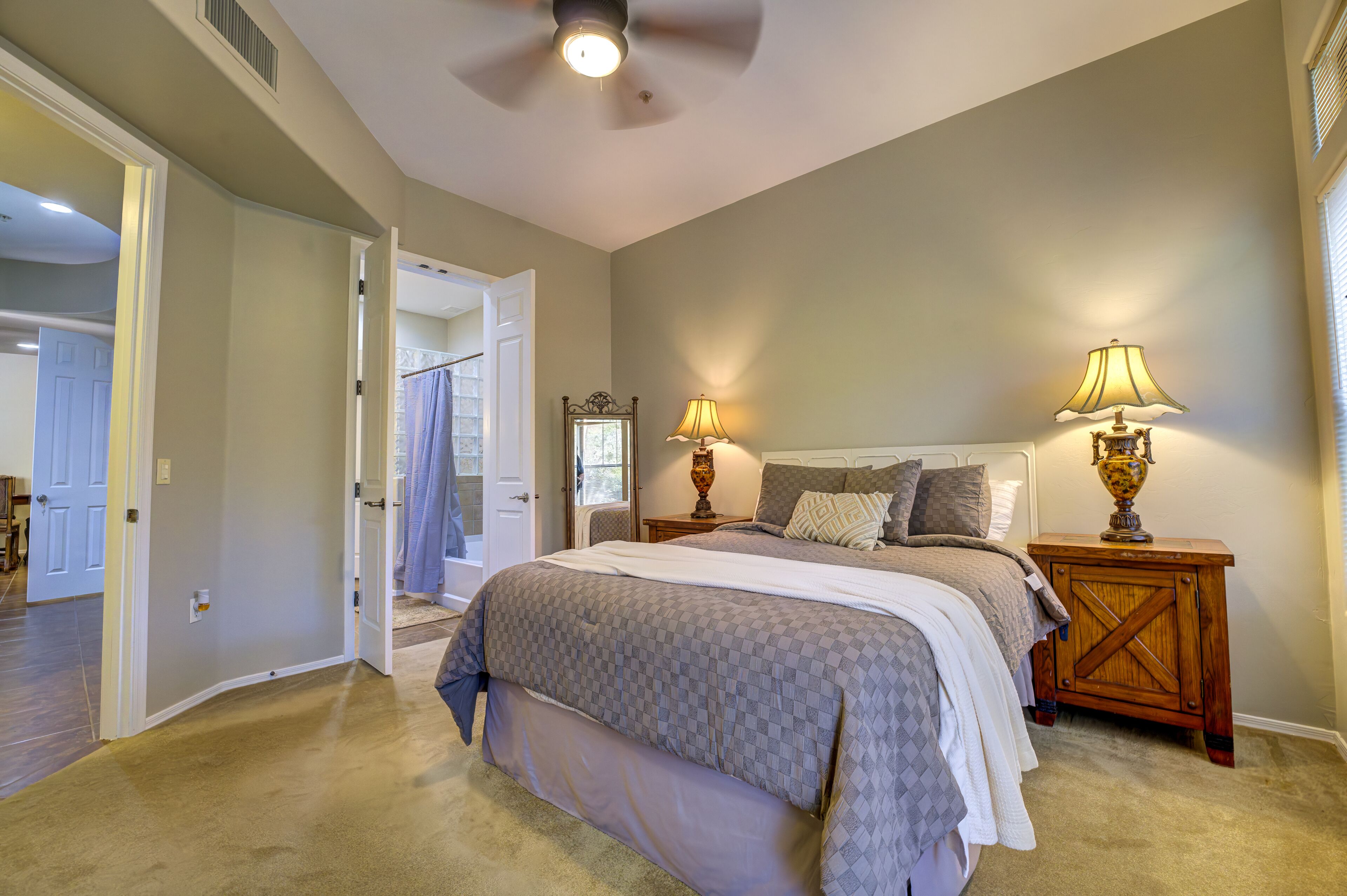 A bedroom with a large bed, two bedside lamps, a ceiling fan, and a view of the bathroom through an open door.