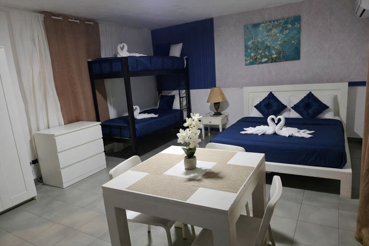 A hotel room with a bunk bed, a single bed, a table with a vase of flowers, and a painting on the wall.