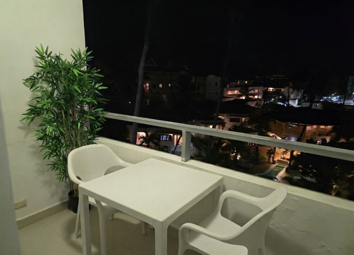 A balcony with a table and chairs, a potted plant, and a view of buildings at night.