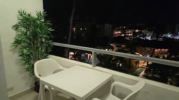 A balcony with a table and chairs, a potted plant, and a view of buildings at night.