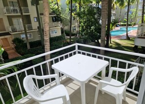 A balcony with a white table and two chairs, overlooking a swimming pool and a building with balconies. - Corales Ducassi Rooftop pool & tropical Caribbean sun (Punta Cana)