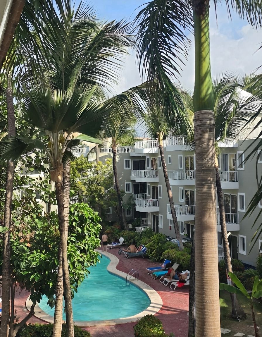 A pool area with lounge chairs and palm trees, surrounded by a multi-story building with balconies.