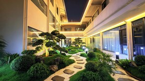 Corredor de hotel bem iluminado, com um caminho de pedra que leva à entrada, cercado por sebes e pequenas árvores bem aparadas.