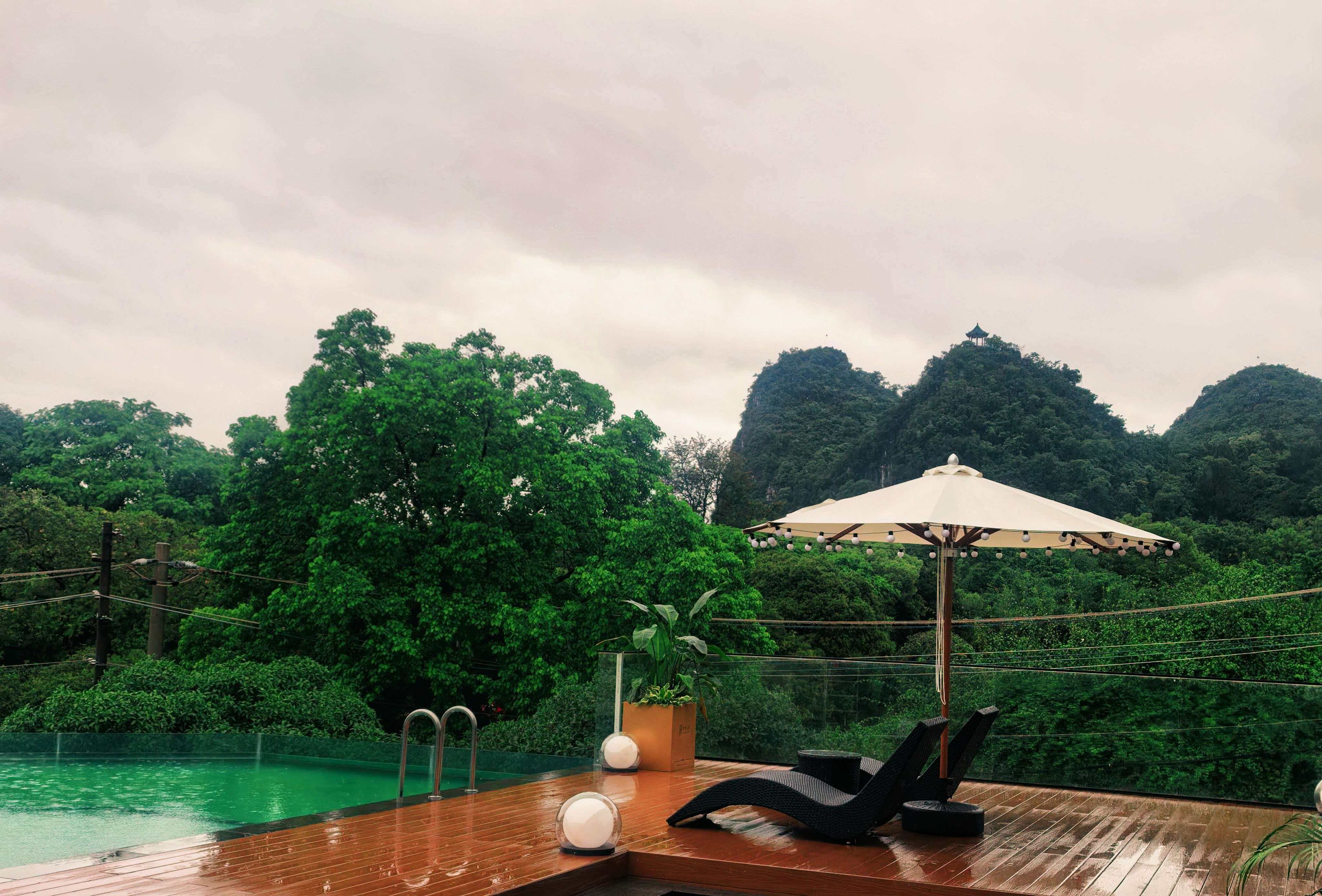 Una terraza de madera con piscina, una silla de descanso, una planta en maceta y una sombrilla.