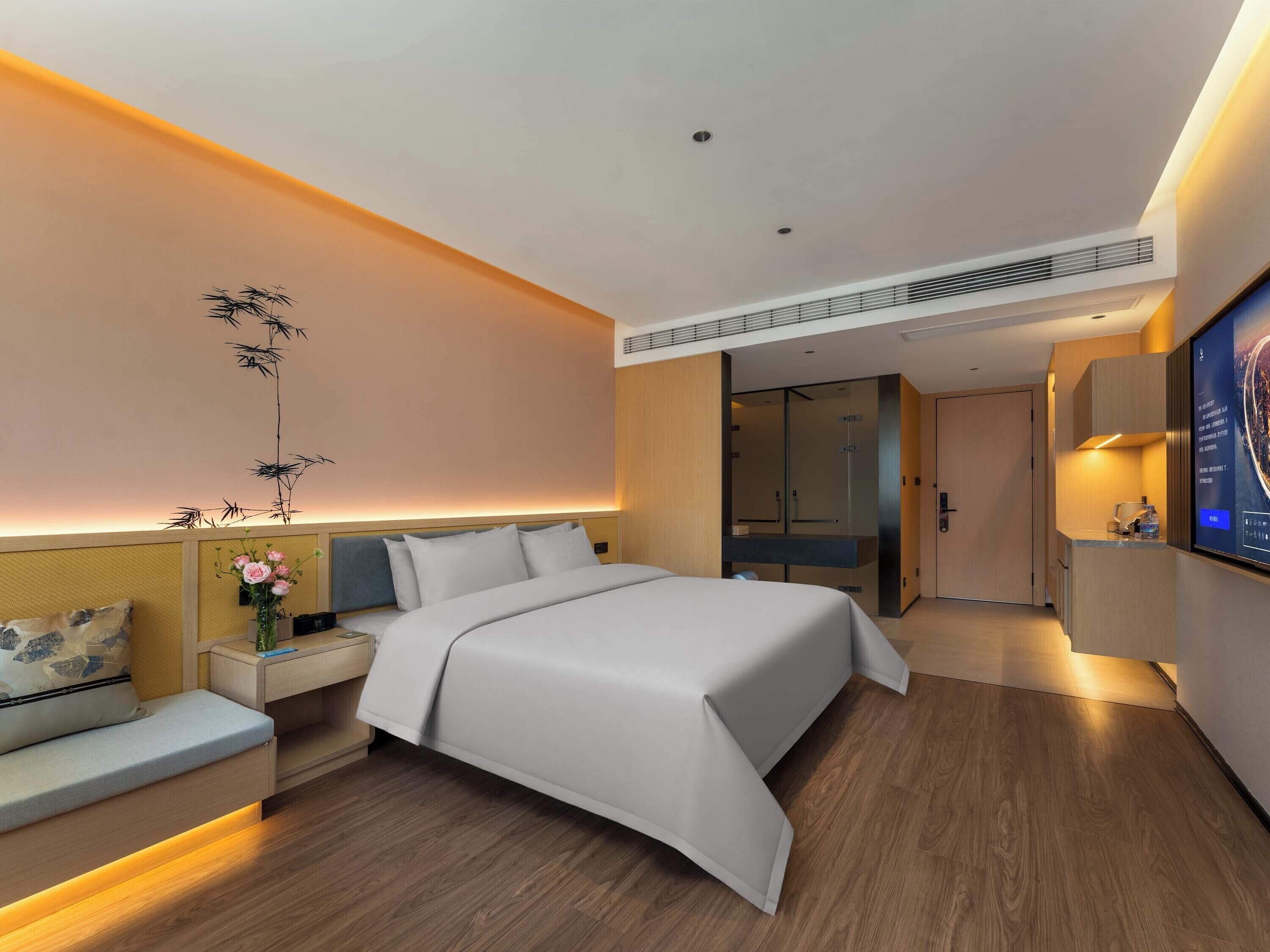 Quarto de hotel moderno com cama grande, mesas de cabeceira, área de estar, televisão presa à parede e guarda-roupa.