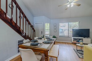 Comedor con mesa de madera dispuesta para cuatro, una silla blanca, un banco, un ventilador de techo y un televisor sobre un mueble.