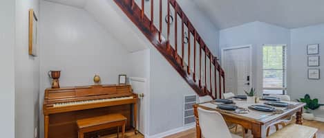 A dining area with a wooden table set for four, a piano, and a staircase with a wooden railing.