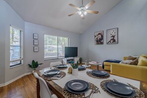 Un comedor con una mesa lista para cuatro personas, un televisor, un sofá y obras de arte en las paredes.