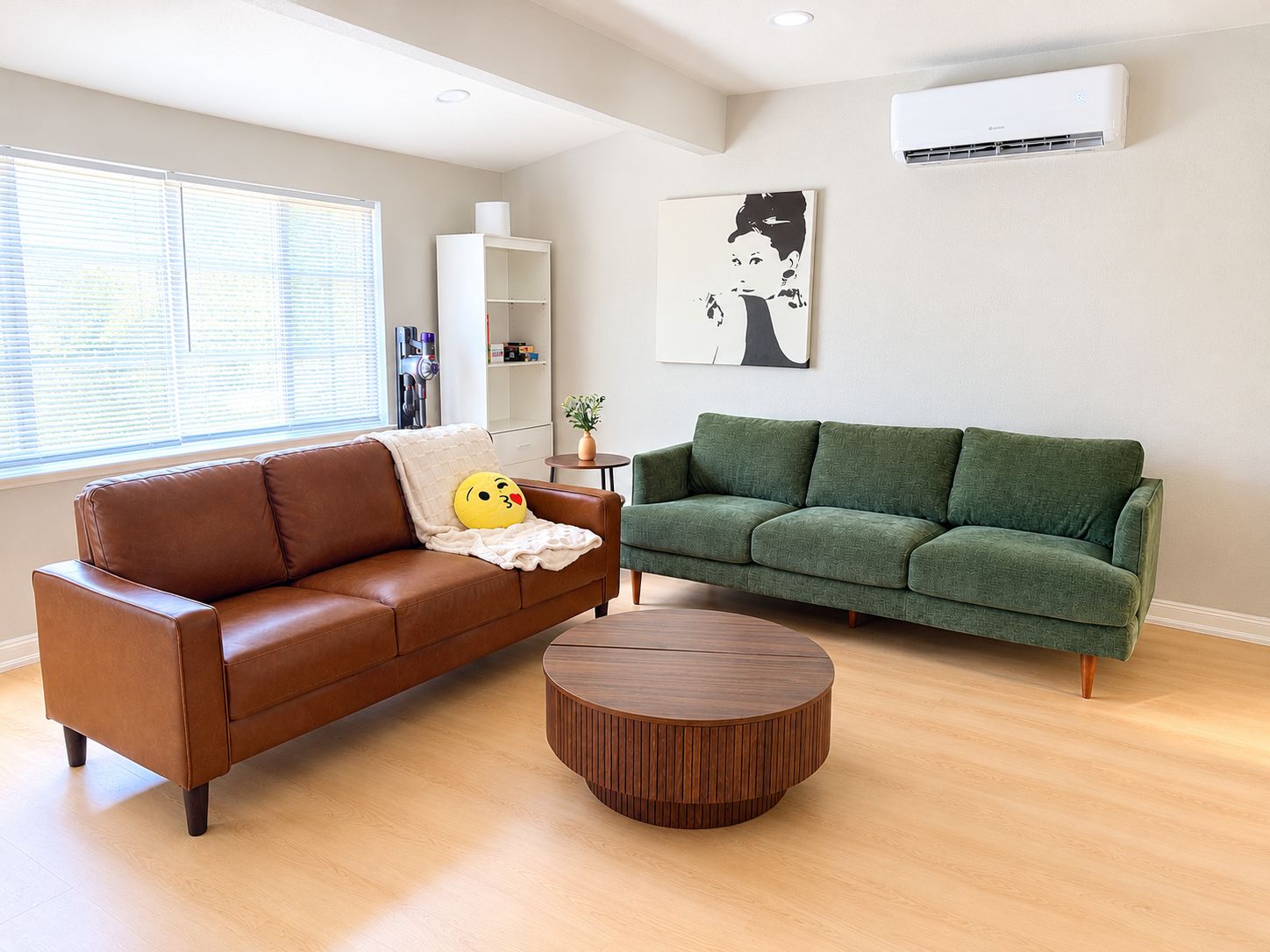 Una sala de estar con un sofá de cuero marrón, un sofá verde, una mesa de centro redonda de madera y un aire acondicionado montado en la pared.