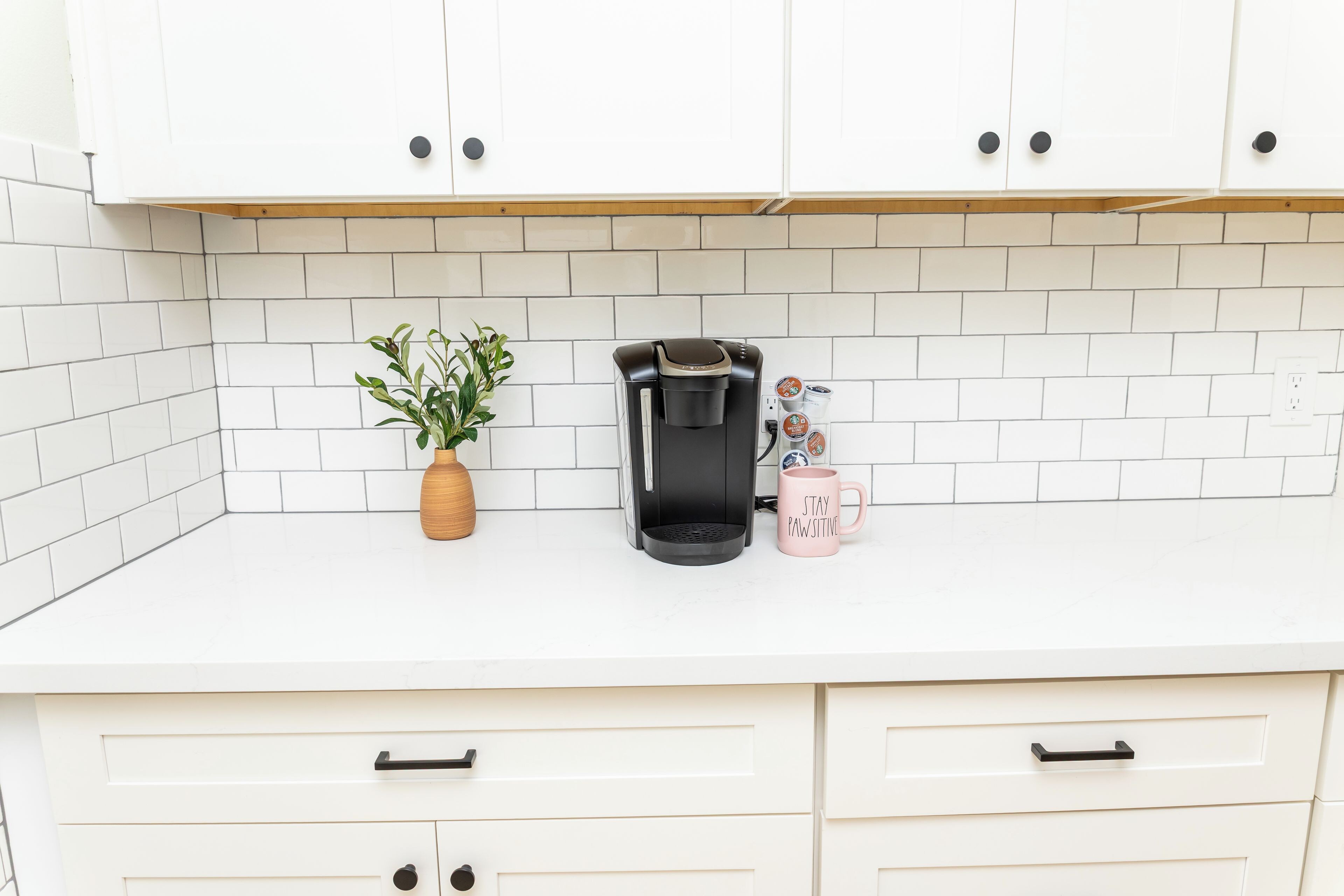 A kitchen with white cabinets, a coffee maker, a plant, and a mug with text.
