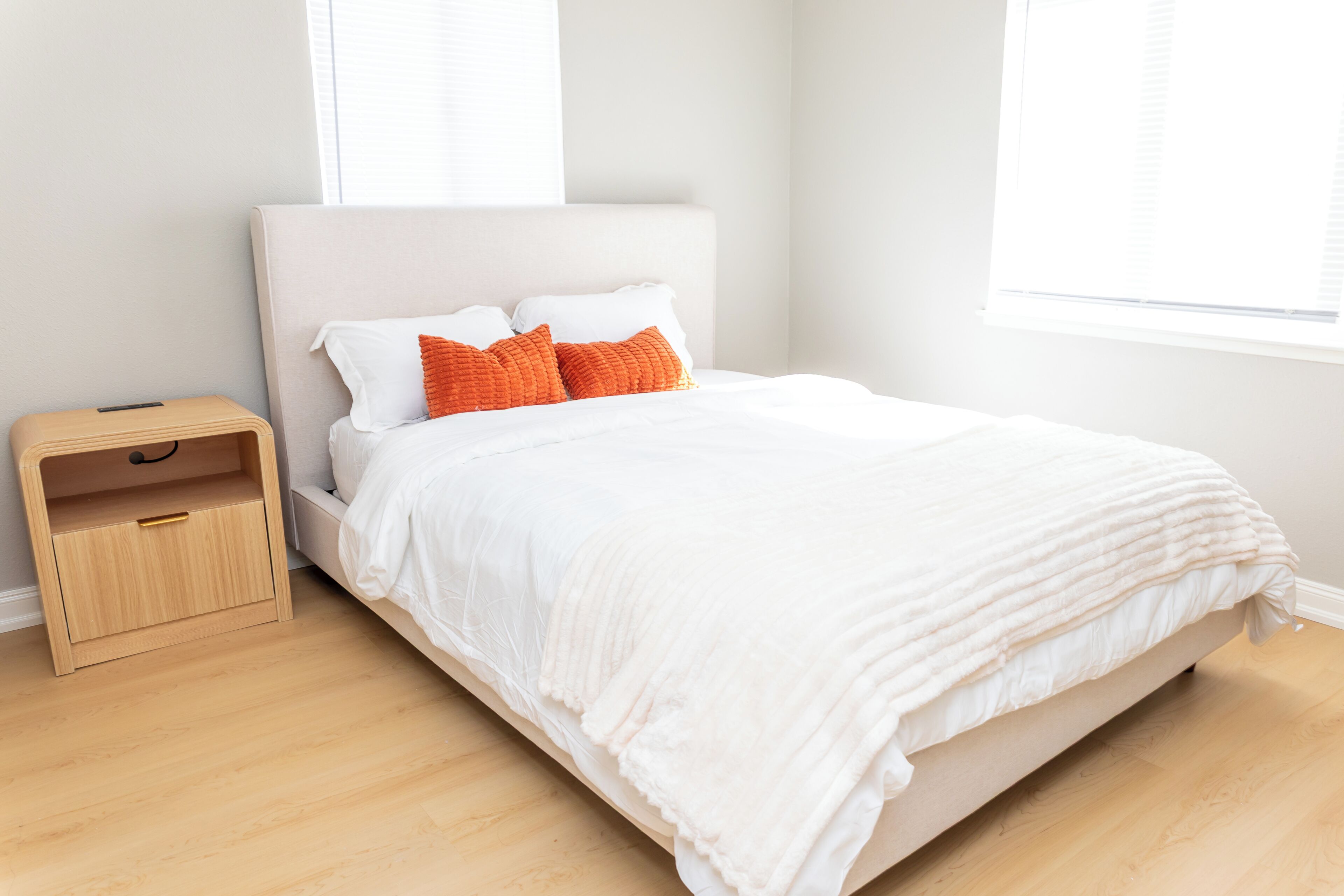 A neatly made bed with white bedding and two orange pillows, a small wooden nightstand with a drawer, and a window with blinds.