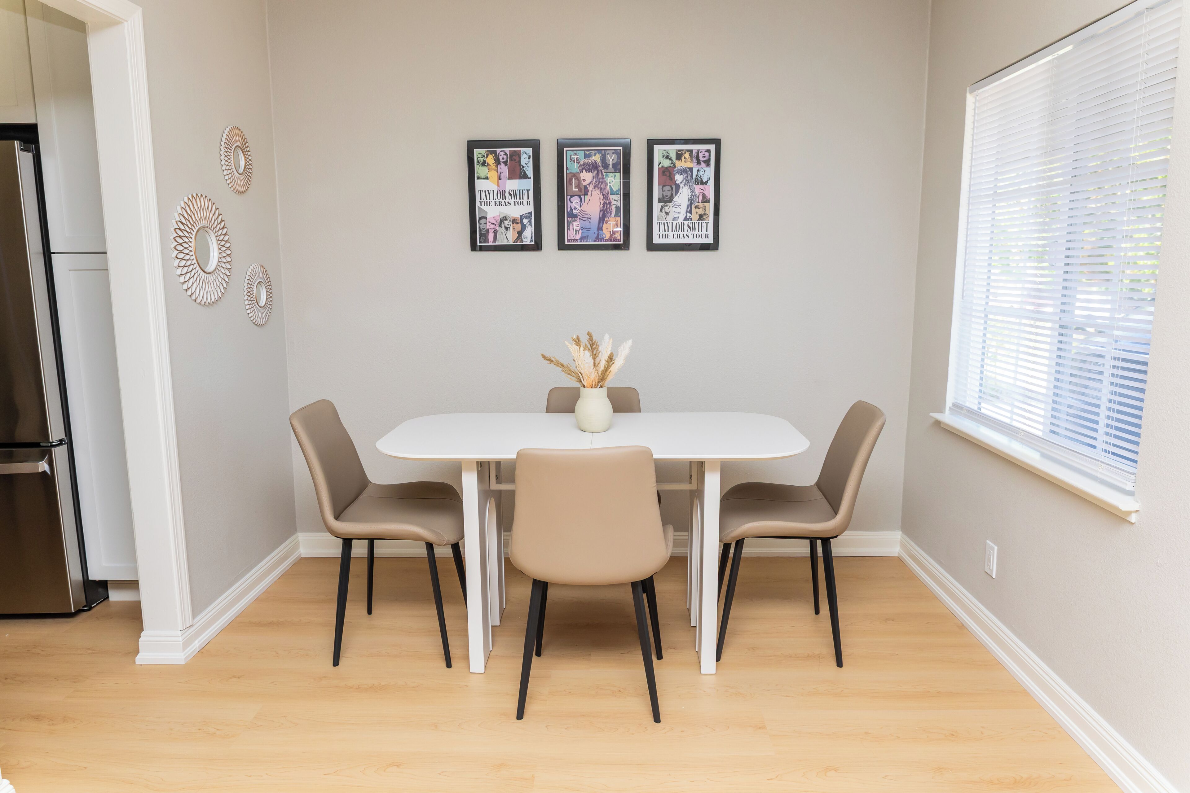A dining area with a white table and four chairs, a vase with dried plants, and three framed posters on the wall.