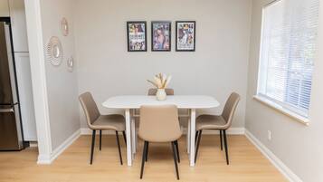 A dining area with a white table and four chairs, a vase with dried plants, and three framed posters on the wall.