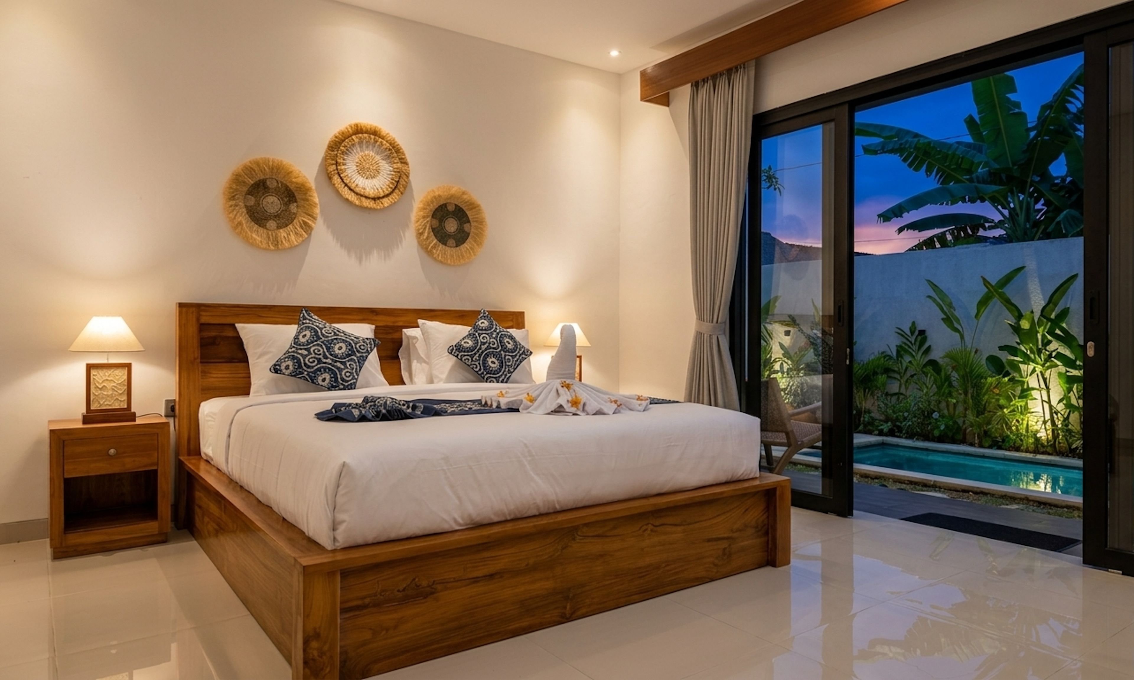 A bedroom with a wooden bed, a nightstand with a lamp, and a view of a pool and plants through a glass door.