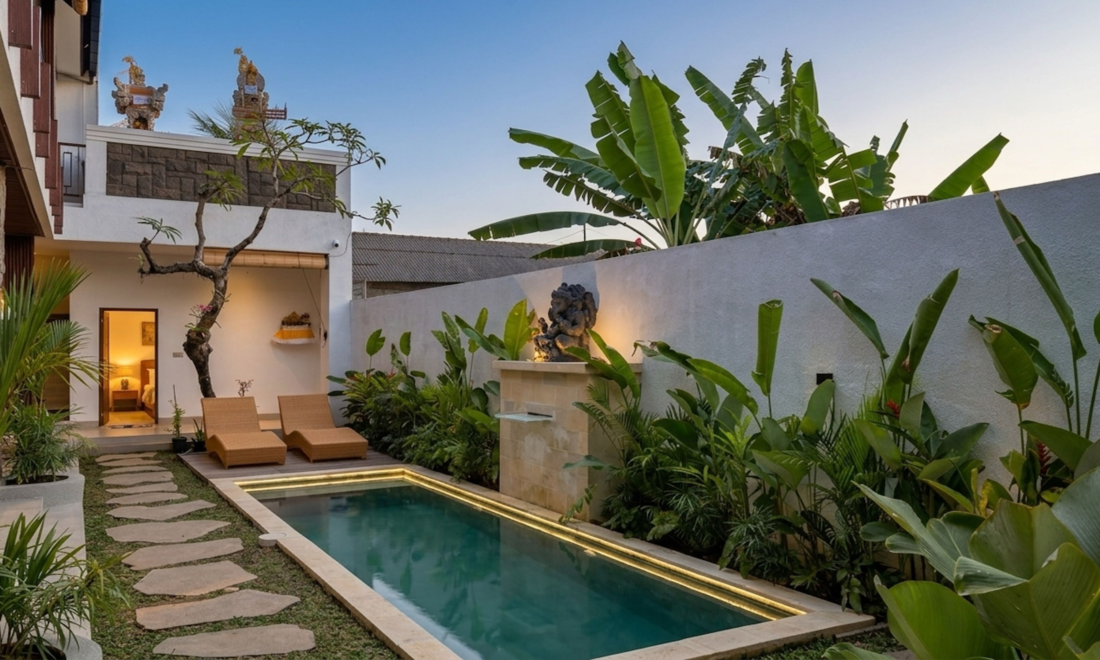 A modern outdoor pool area with a stone pathway leading to a seating area, surrounded by lush greenery and a white wall.
