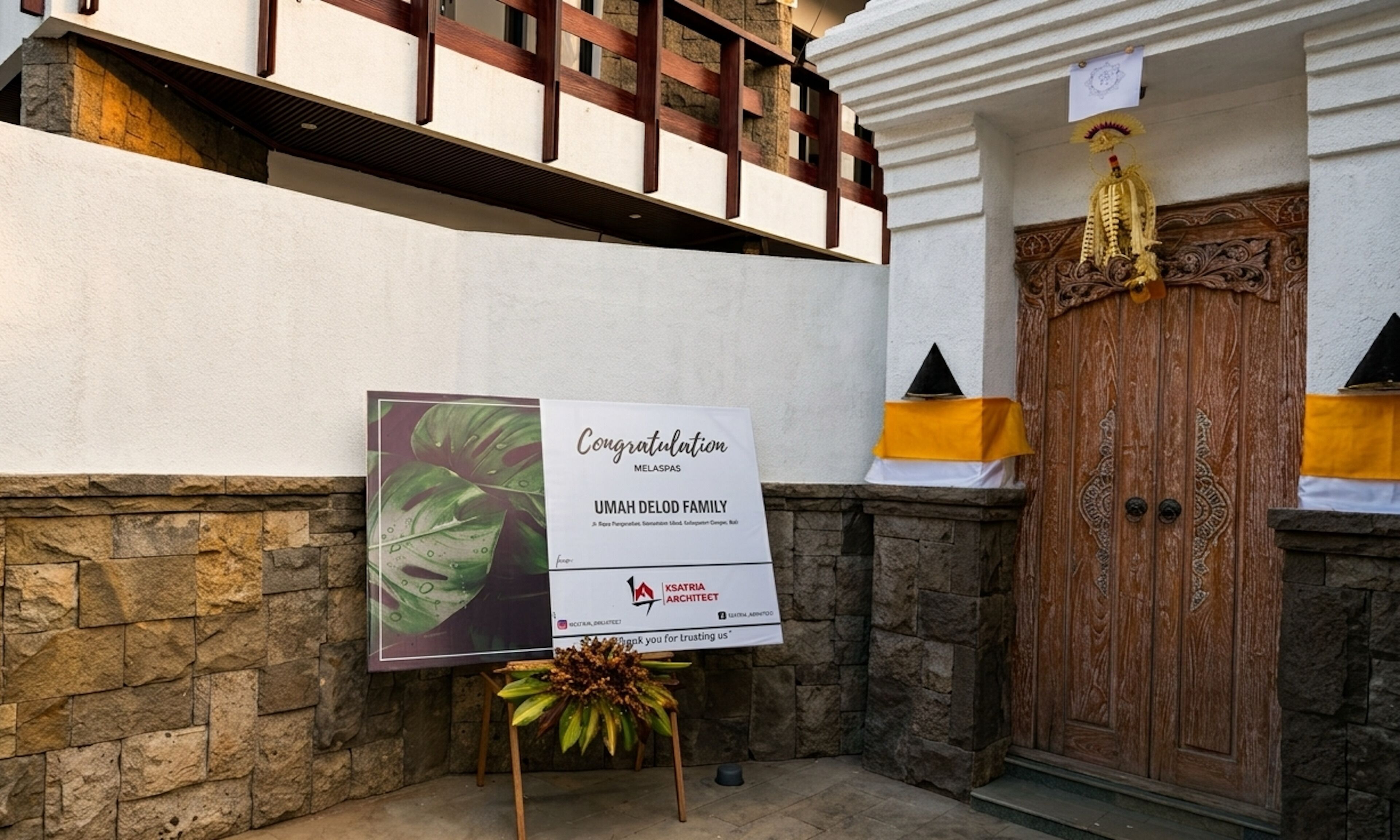 Un letrero de felicitación para la familia UMAH DELOD, colocado sobre un atril con una planta, frente a un muro de piedra y una puerta de madera con detalles decorativos.