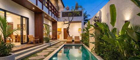 A modern building with a swimming pool, outdoor seating, and tropical plants.