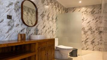 Un baño con pared de mármol, tocador de madera, lavamanos, inodoro y cabina de ducha con mampara de vidrio.