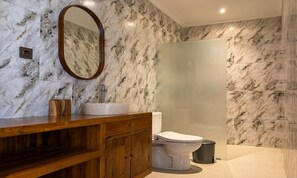 Un baño con pared de mármol, tocador de madera, lavamanos, inodoro y cabina de ducha con mampara de vidrio.