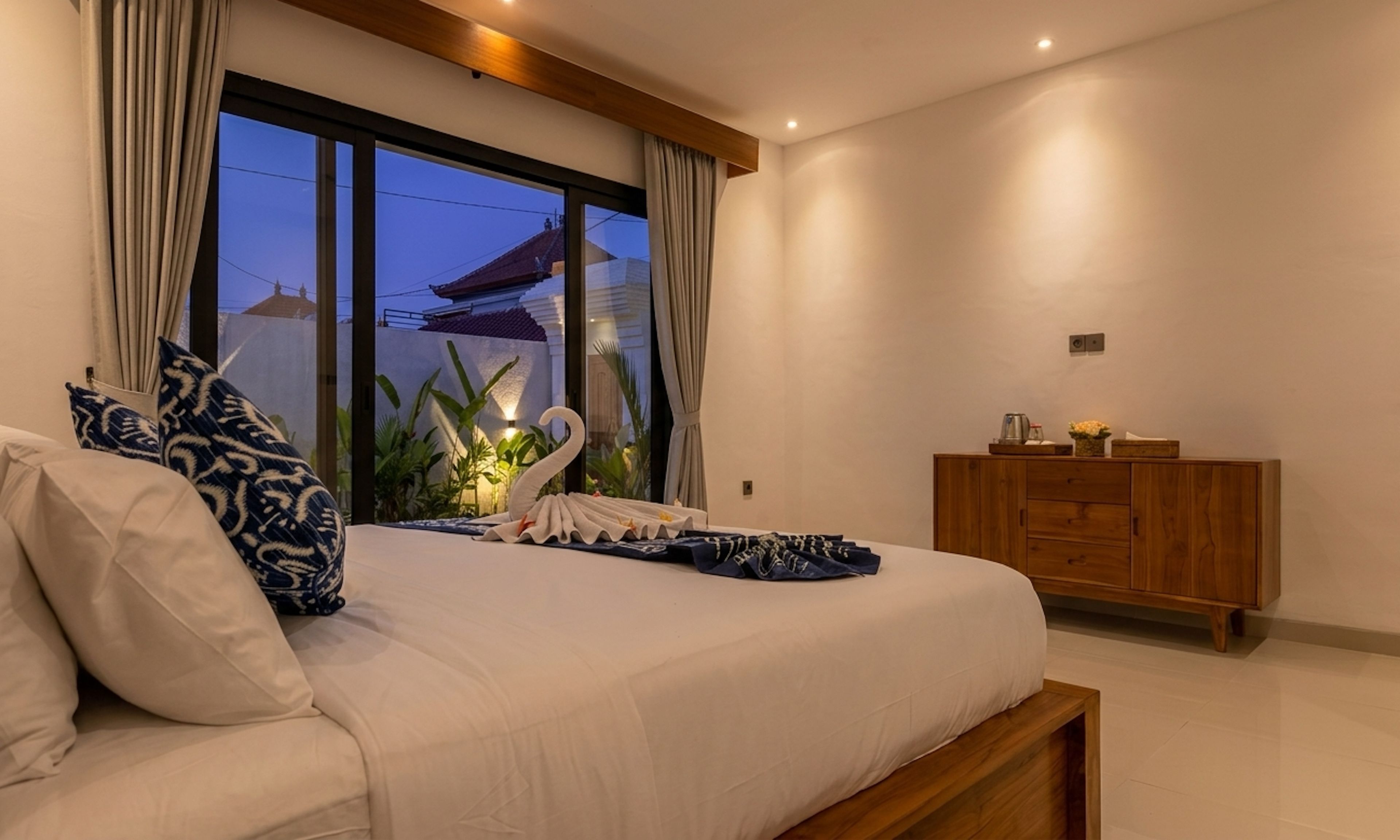 A bedroom with a large bed, curtains, a wooden dresser, and a view of buildings outside.