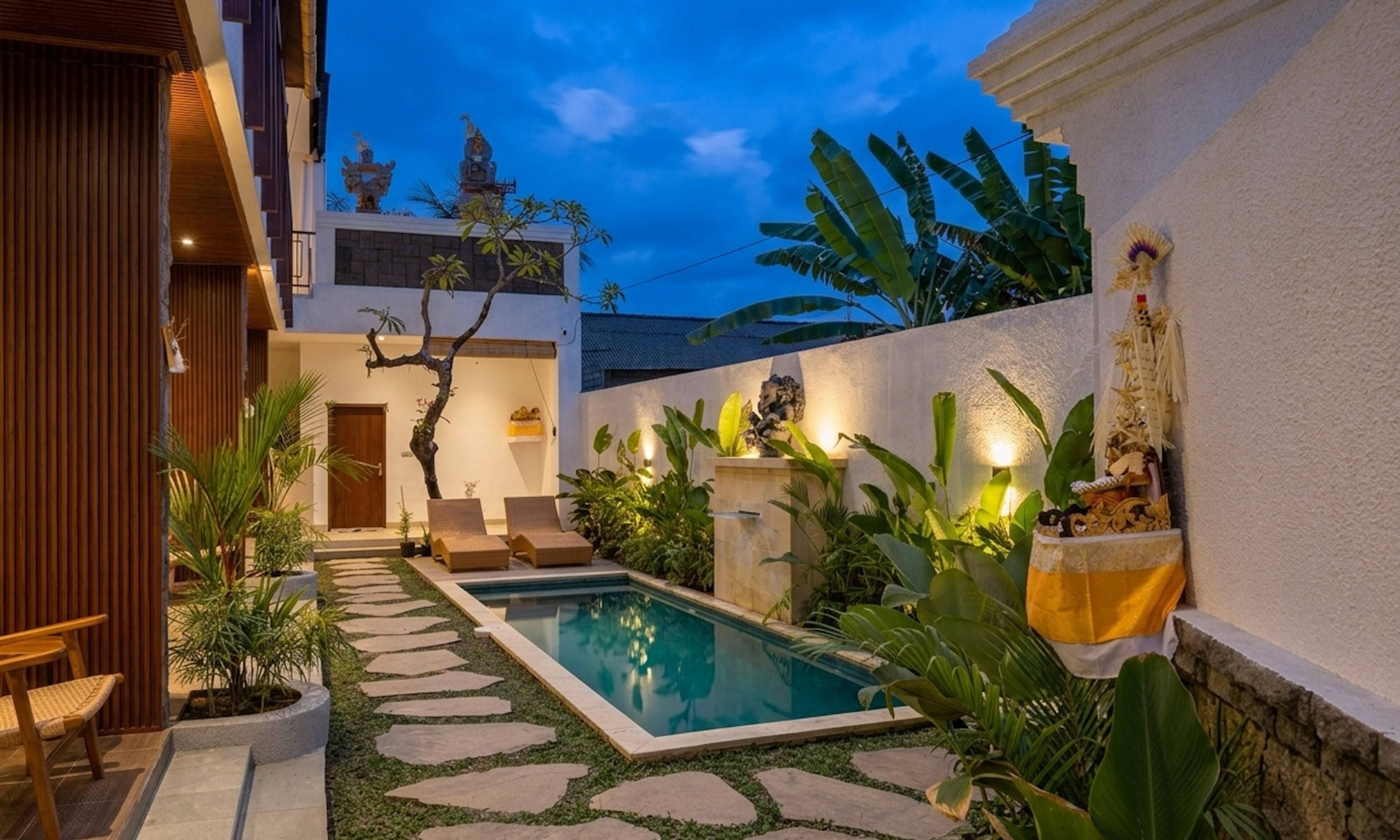 A pool area with stone steps leading to a pool, surrounded by tropical plants and a white wall with decorative elements.