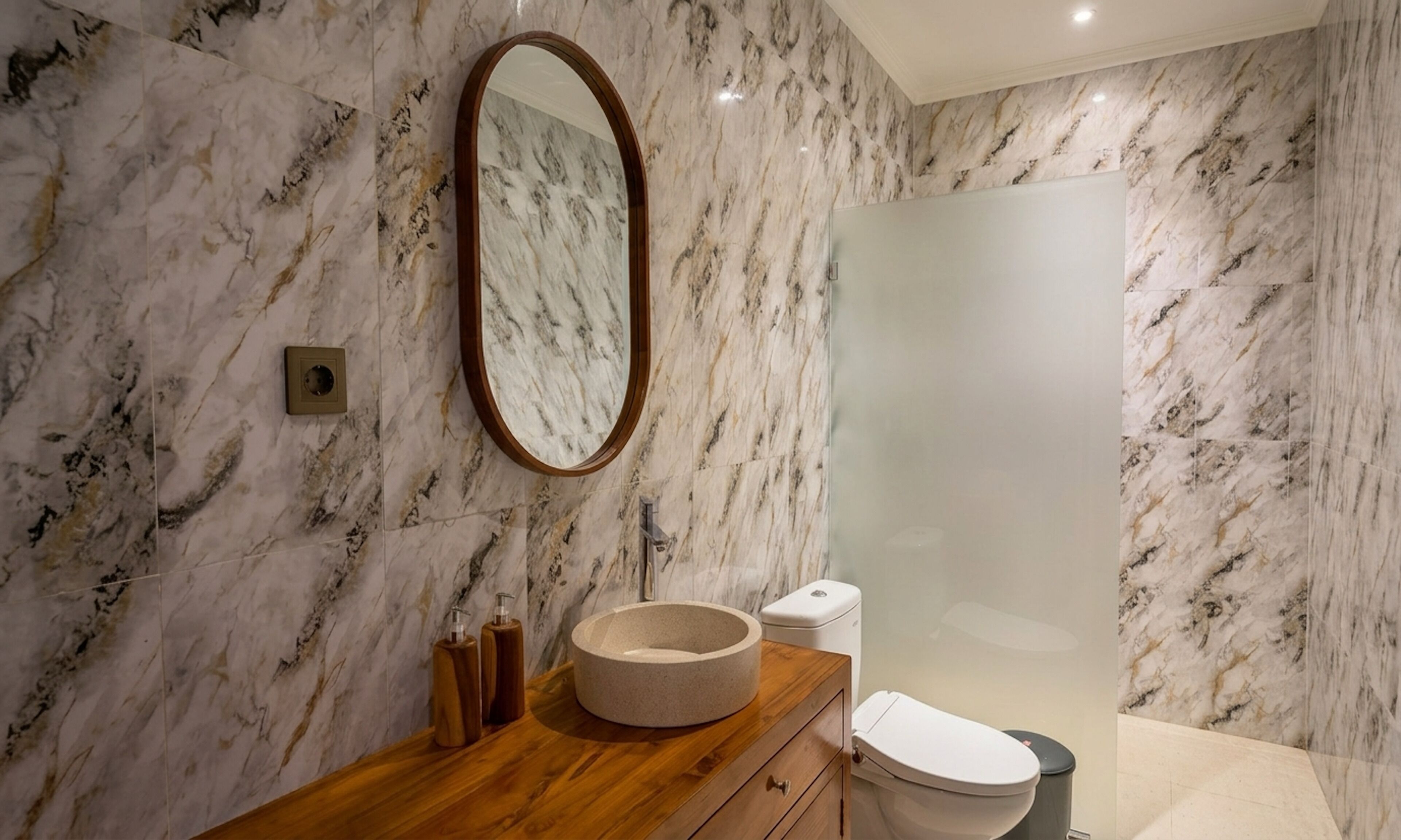 A bathroom with a marble wall, a wooden vanity, a round basin, a toilet, and a mirror.