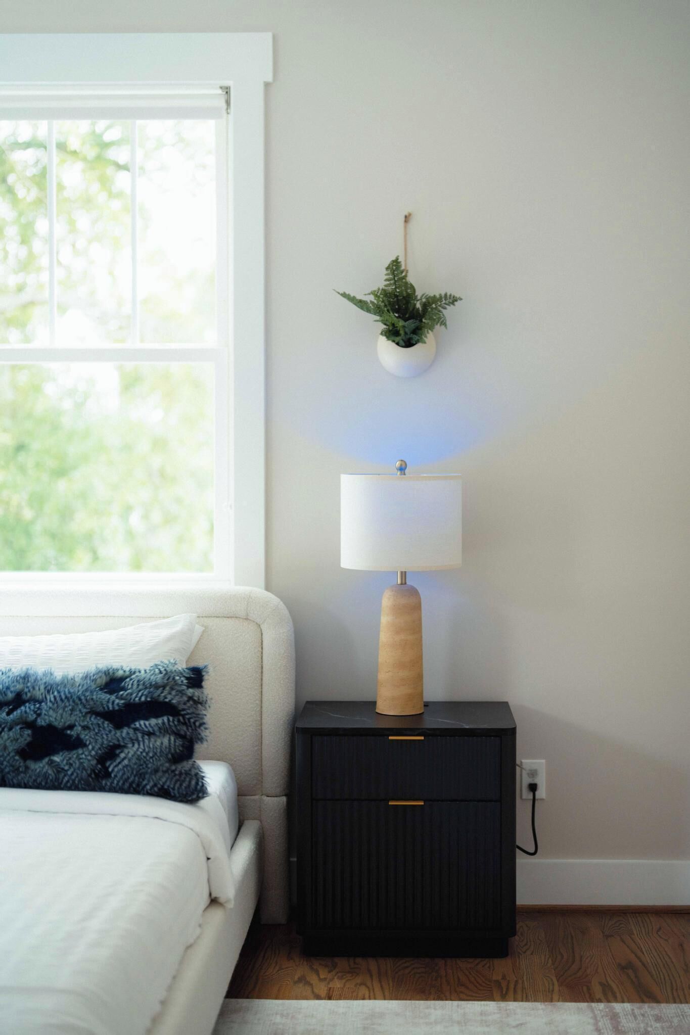 A bedroom with a bed, a bedside table with a lamp, a wall-mounted plant, and a window with daylight.