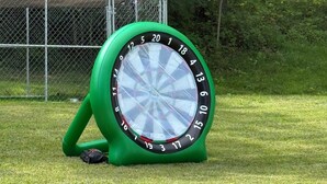 Un bersaglio da freccette con supporto verde, posizionato su un prato con una recinzione a maglie di filo sullo sfondo.