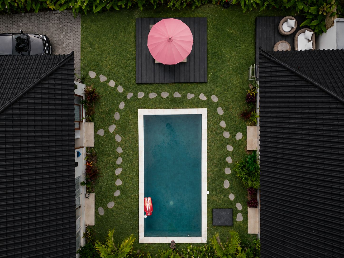 Une piscine rectangulaire entourée d’un chemin de pierre, avec un parasol rose et une chaise longue rouge.