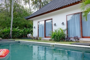 A swimming pool with a tiled deck, surrounded by lush greenery and a modern house with large windows and a tiled roof.