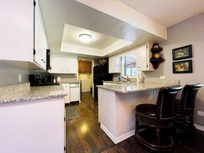 A compact kitchen with white cabinets, a granite countertop, a built-in island with bar stools, a refrigerator, and a clock on the wall. - Legacy Homestead Cabin, White Mountains AZ (Navajo County)