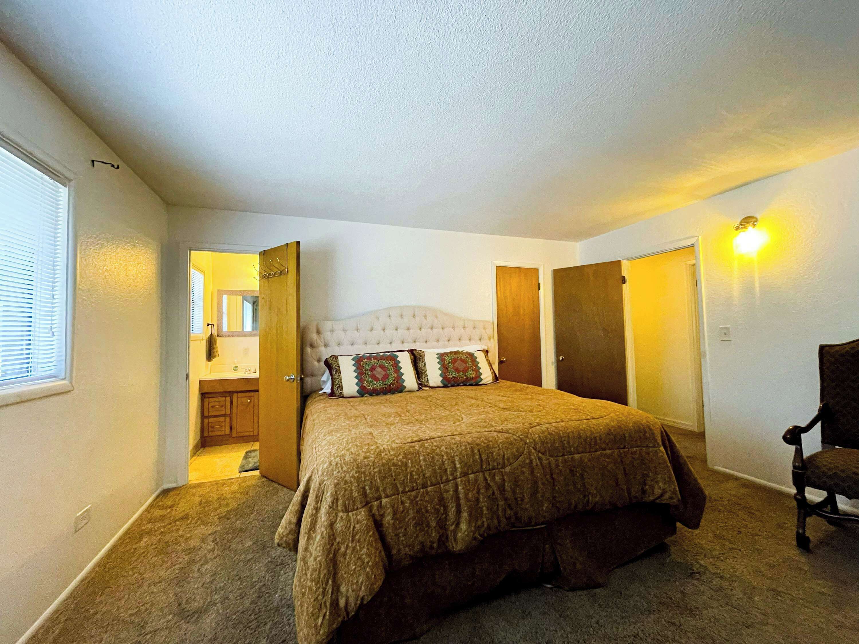 Une chambre à coucher avec un grand lit, une chaise, une armoire et une salle de bain visible à travers une porte ouverte.