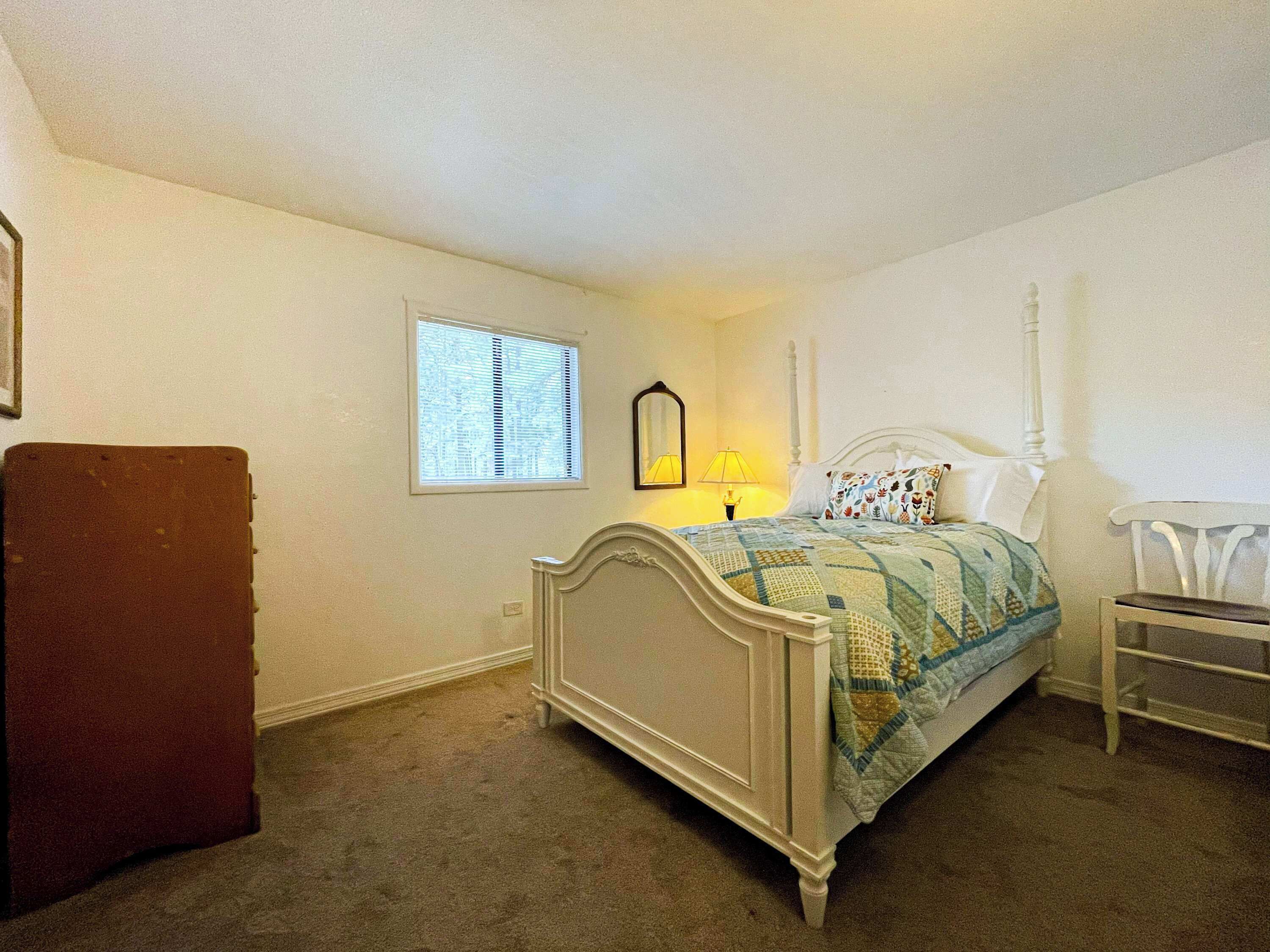 Ein Schlafzimmer mit einem Bett, einem Sessel, einer Kommode, einem Fenster mit Jalousien und einem Spiegel an der Wand.